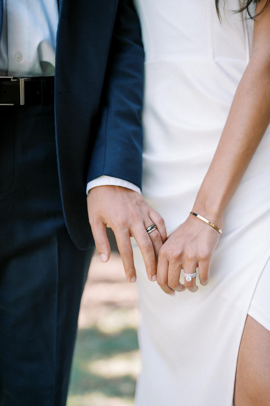 A close-up of a couple holding hands, showcasing wedding bands and elegant attire, with the groom in a navy suit and the bride in a white dress, symbolizing love and commitment.