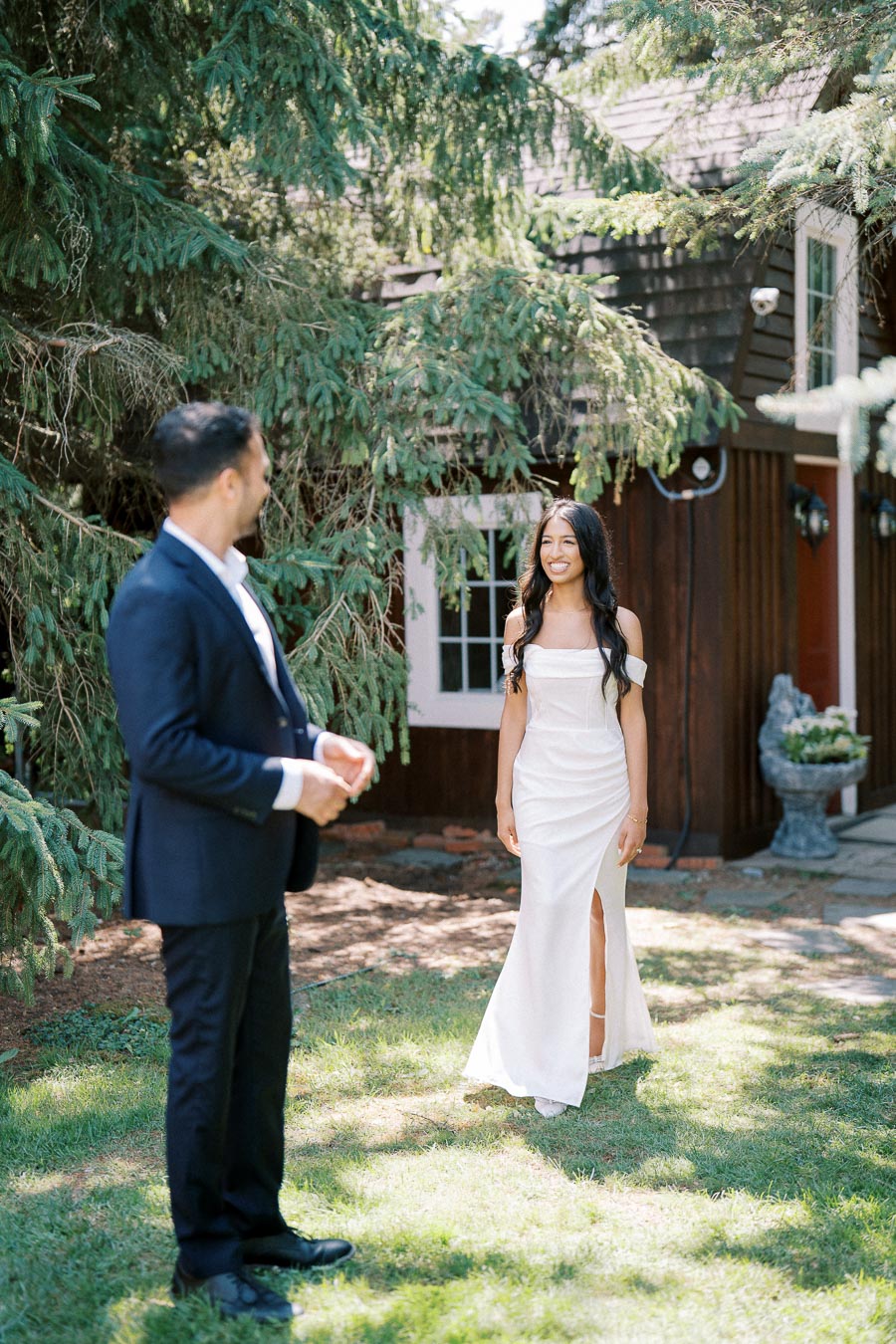 A couple in formal attire standing in a garden setting, with lush greenery and a rustic house in the background. The woman is wearing a white dress and the man is in a dark suit, both smiling and enjoying a sunny day.