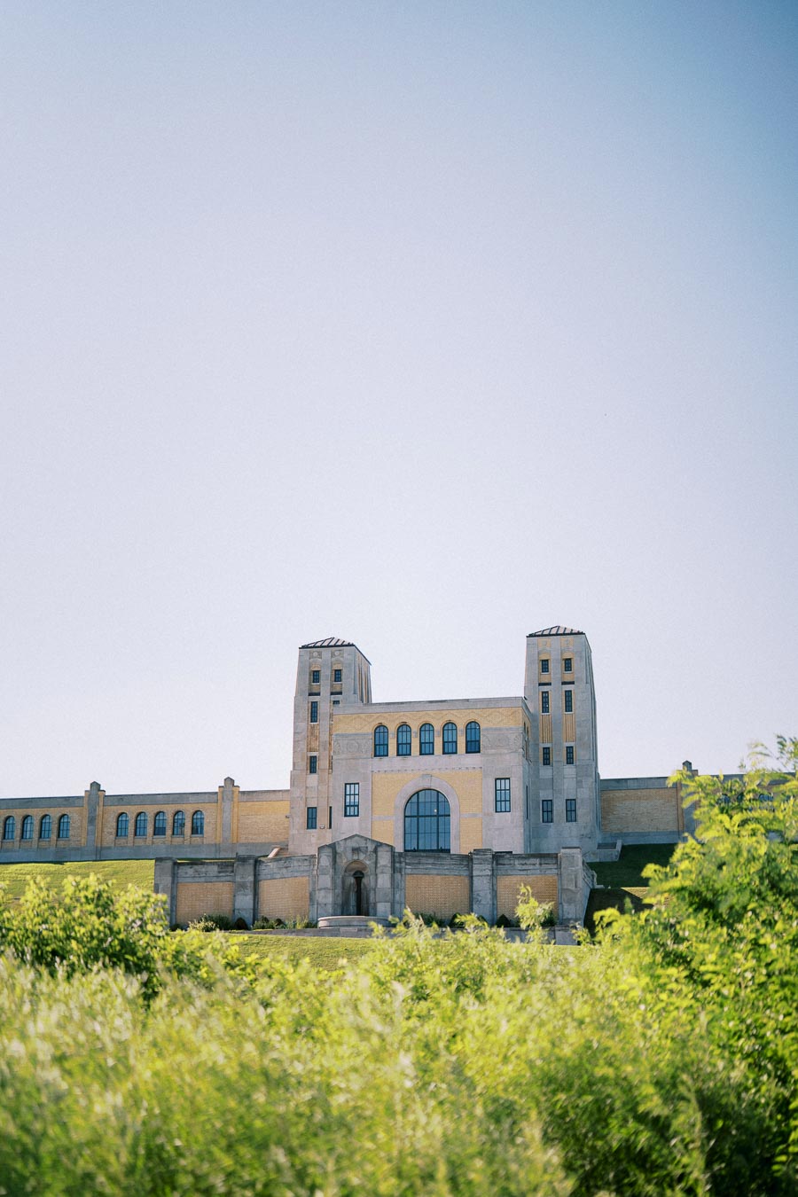 Historic building with two towers and arched windows against a clear blue sky, surrounded by lush greenery.