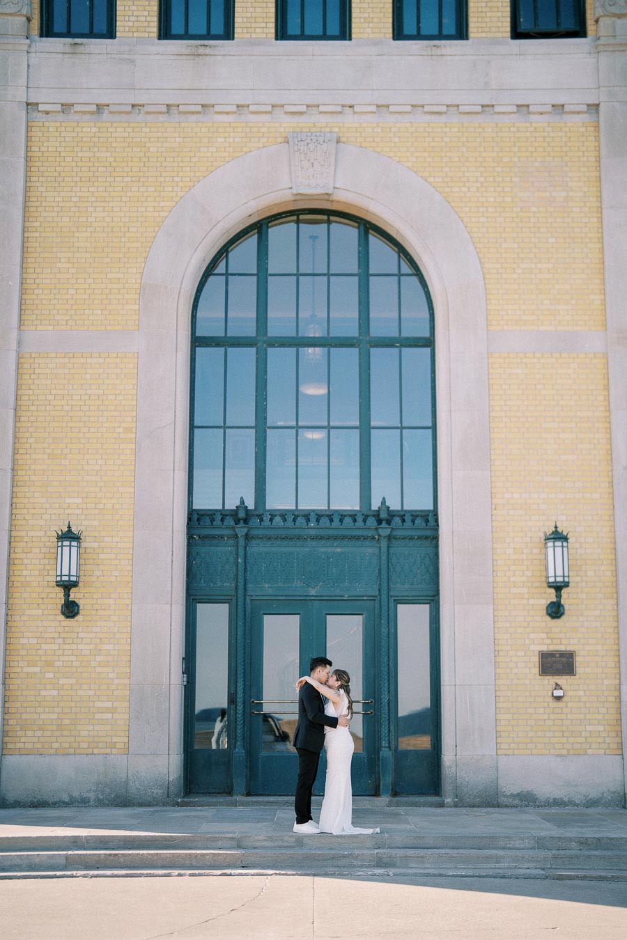 A couple embracing in front of a large, arched door of a historic yellow brick building.