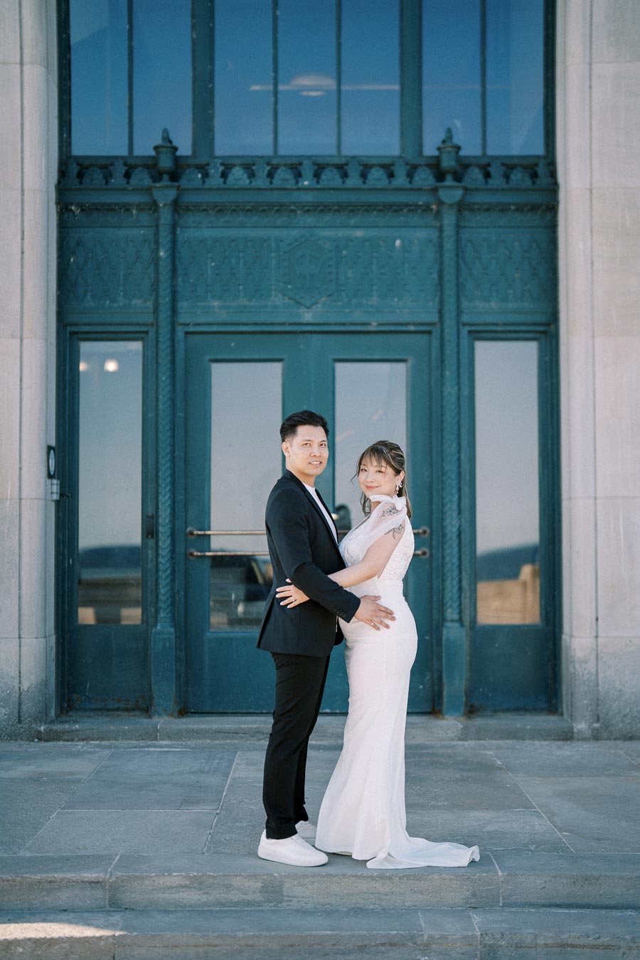 A couple posing in elegant wedding attire, with the bride in a classic white gown and the groom in a black suit, standing in front of a large, ornate blue door. The photo captures a serene moment of joy and connection on their special day.