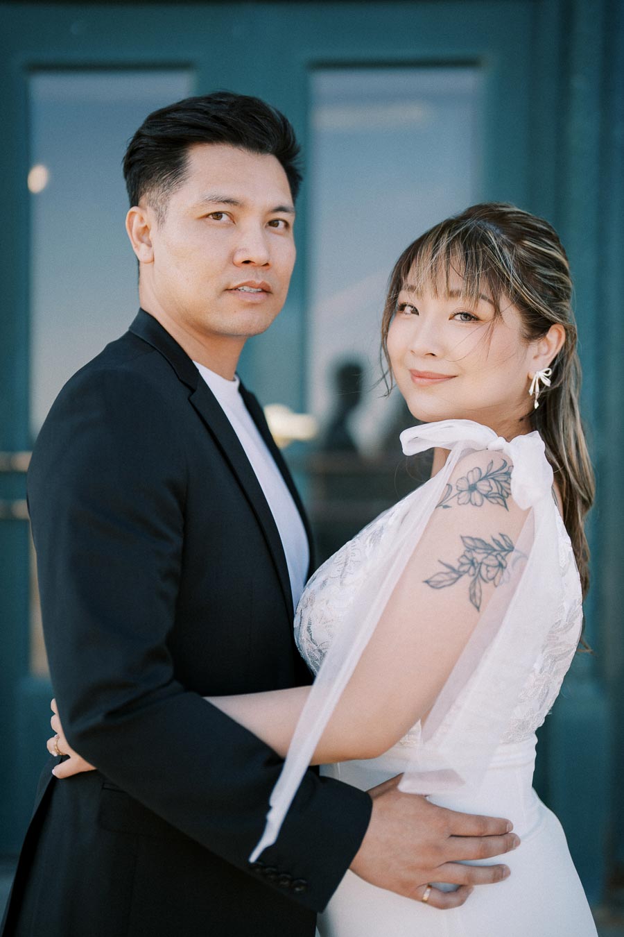 Elegant couple posing in formal attire, woman in white dress with floral tattoo, in front of teal door, displaying wedding bliss and modern romance.