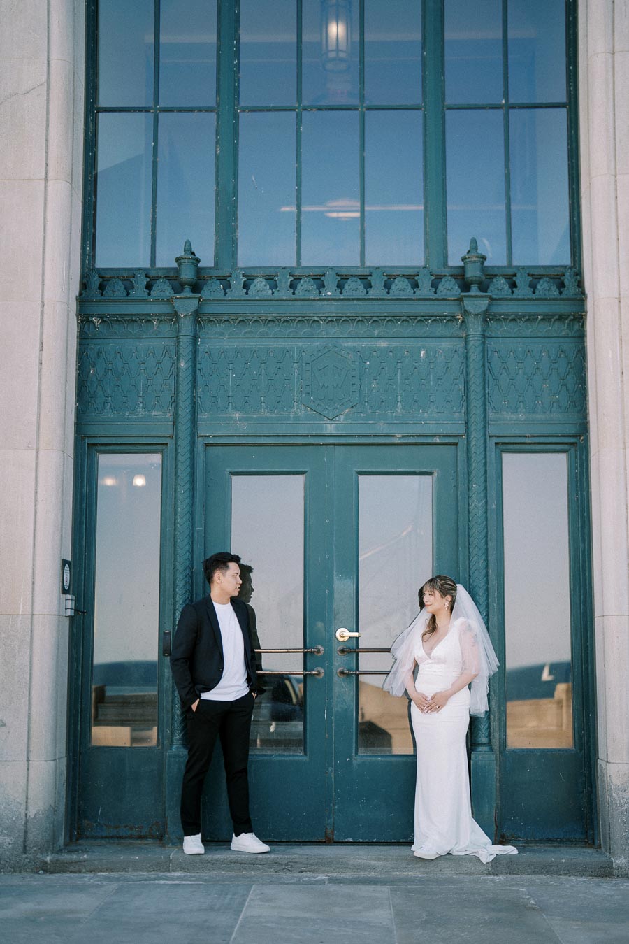 A bride and groom stand facing each other in front of a large blue door with intricate architectural details. The bride wears a white wedding dress and veil, while the groom is in a casual suit with white sneakers.