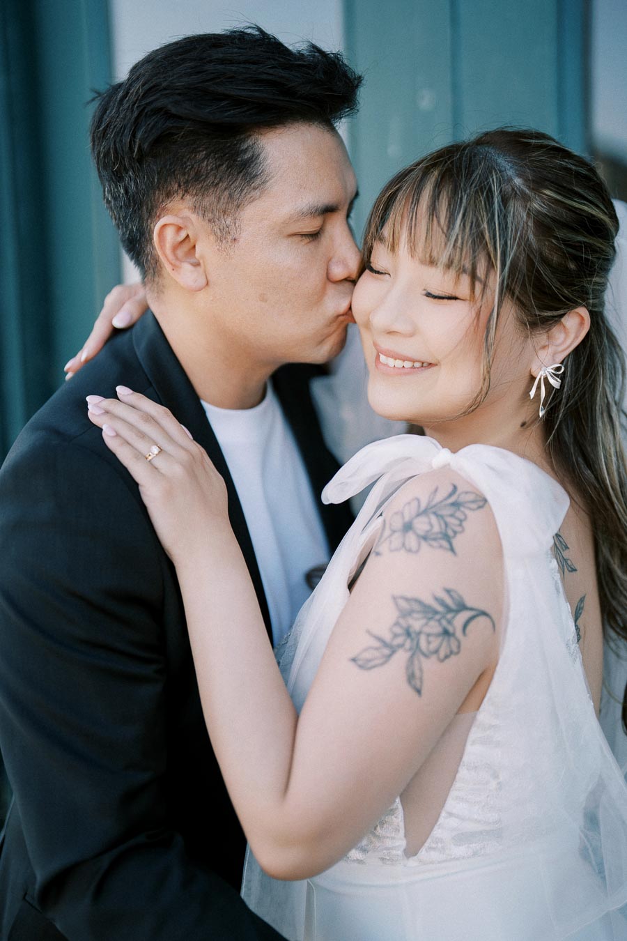 Smiling couple embracing, man in black suit kisses woman's cheek, showcasing her floral tattoo and elegant earrings.
