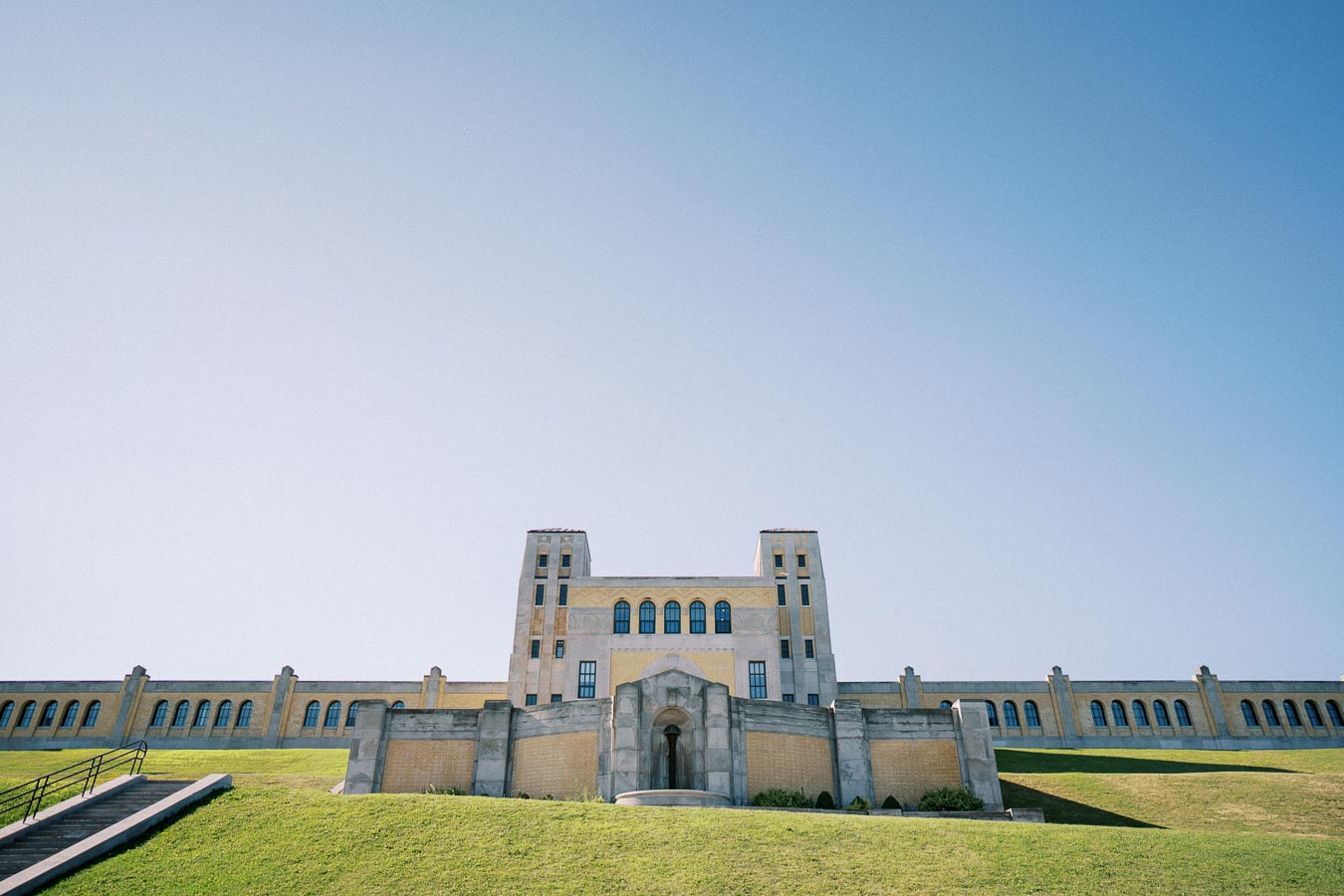 Historic building with a symmetrical facade and arched windows, set against a clear blue sky, viewed from a grassy hill.