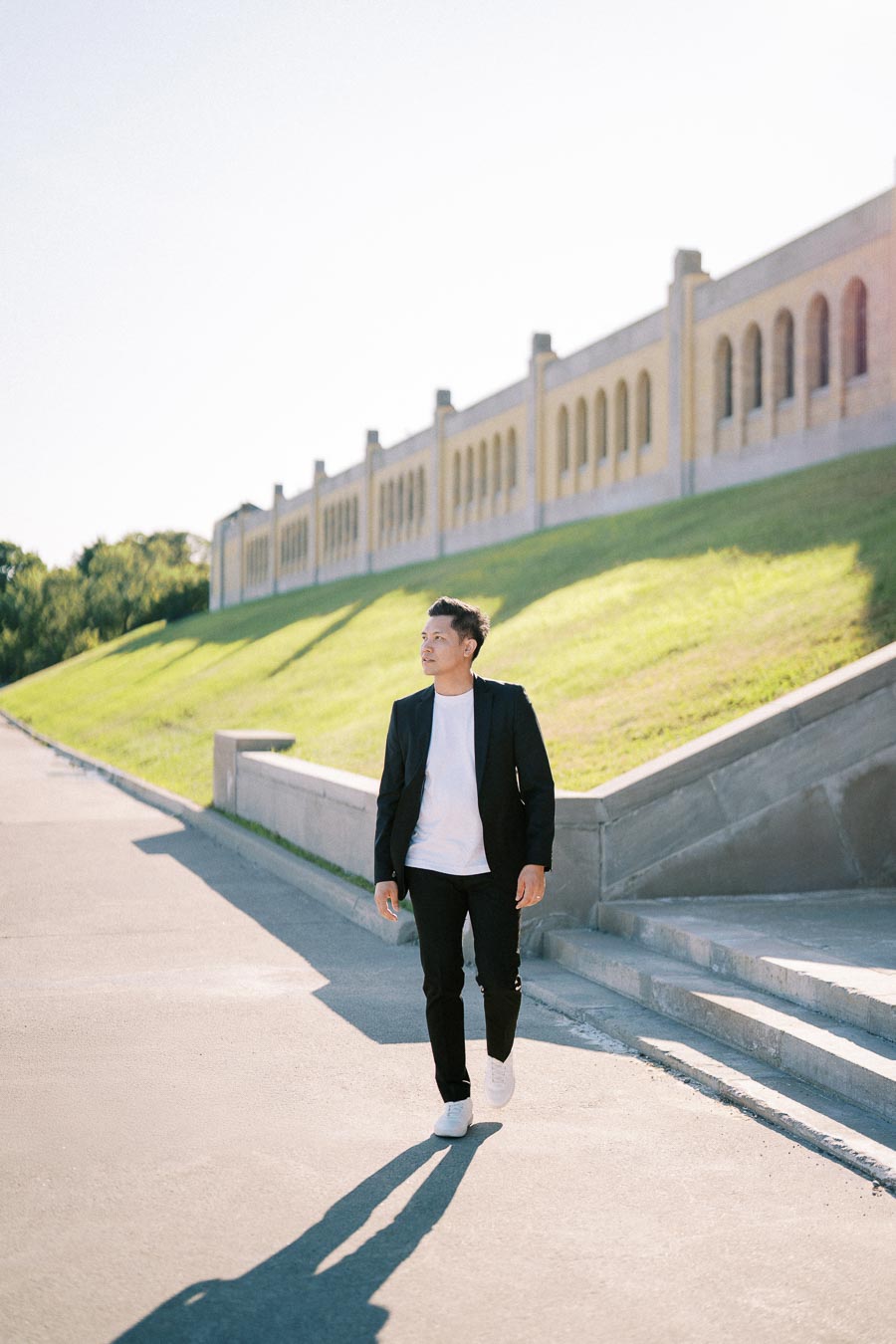 A man in a black suit and white t-shirt walking outdoors on a sunny day next to a historic building with arches and a sloped grassy landscape.