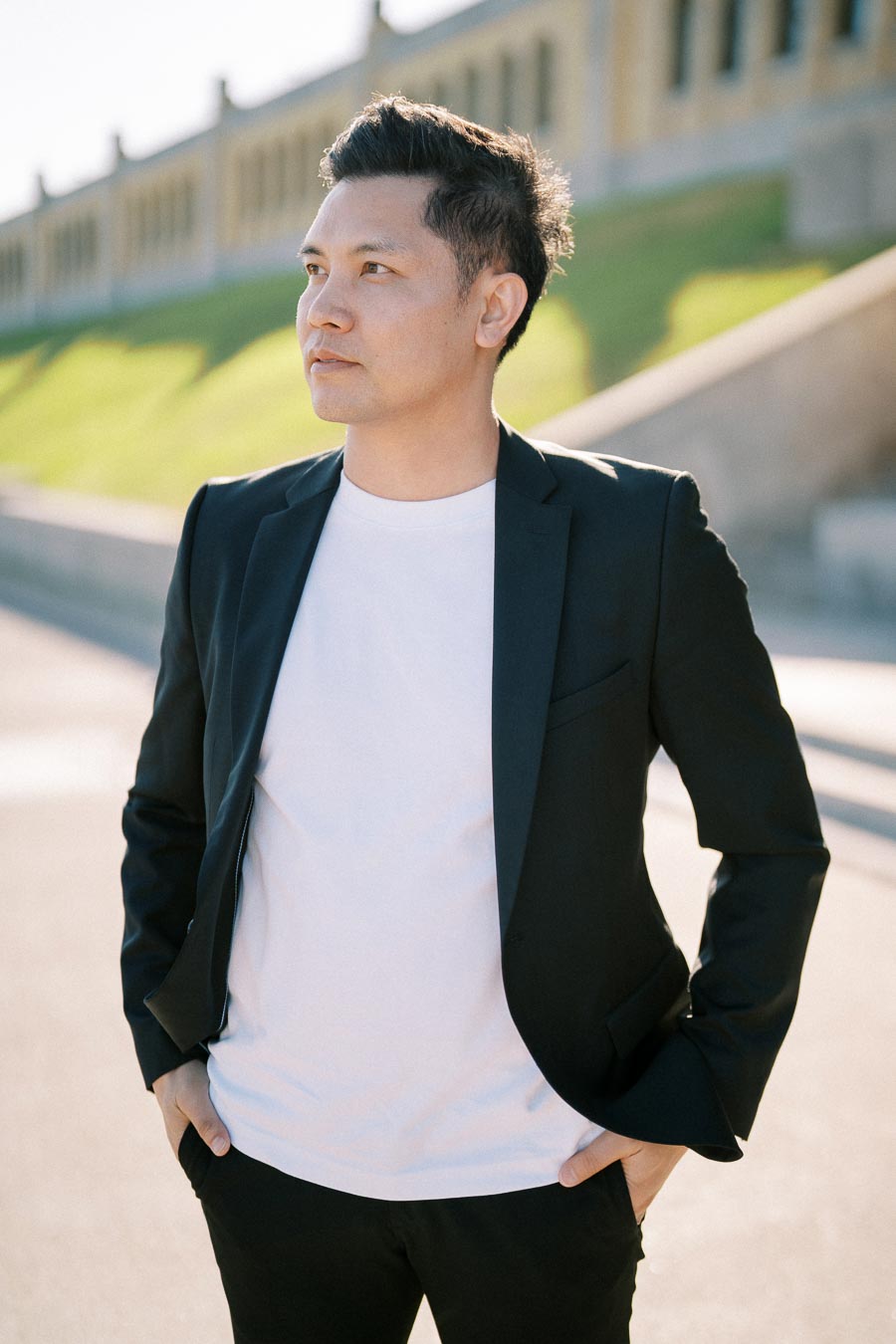 A person wearing a dark blazer and white t-shirt stands confidently outdoors in a sunlit setting, with a blurred background featuring a building and green grass.