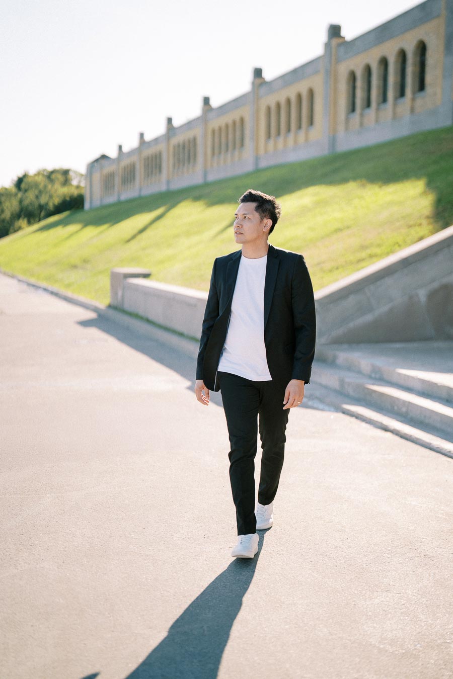 Man in a black suit and white shirt walking outdoors by a historic stone building with arches, under a clear sky.