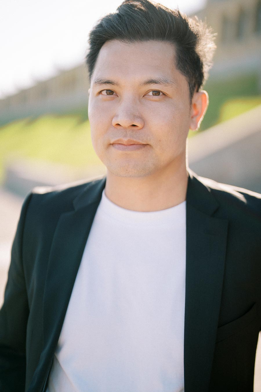 Portrait of a man outdoors wearing a black blazer and white shirt, with blurred grassy background in sunlight.