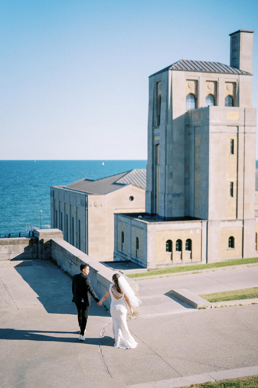 A bride and groom holding hands walk along a pathway with a historic building and scenic ocean view in the background under a clear blue sky.