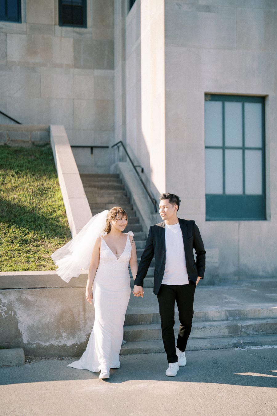 A bride and groom holding hands and smiling while walking outside a stone building and bathed in sunlight, showcasing a beautiful wedding moment.