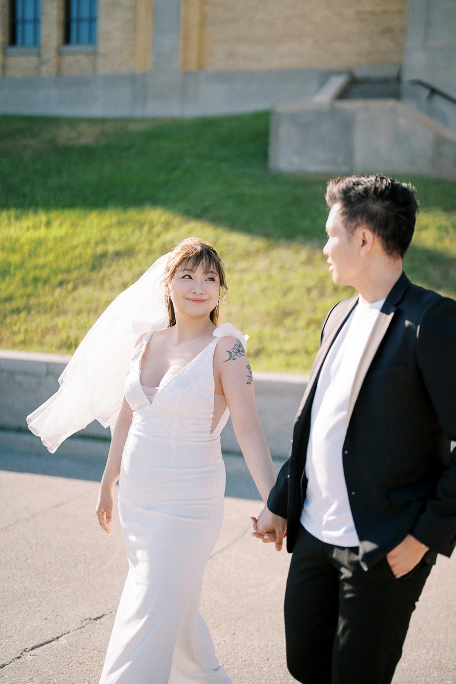 Smiling bride in white dress and veil holding hands with groom in black suit, walking outdoors on a sunny day in front of a stone building.