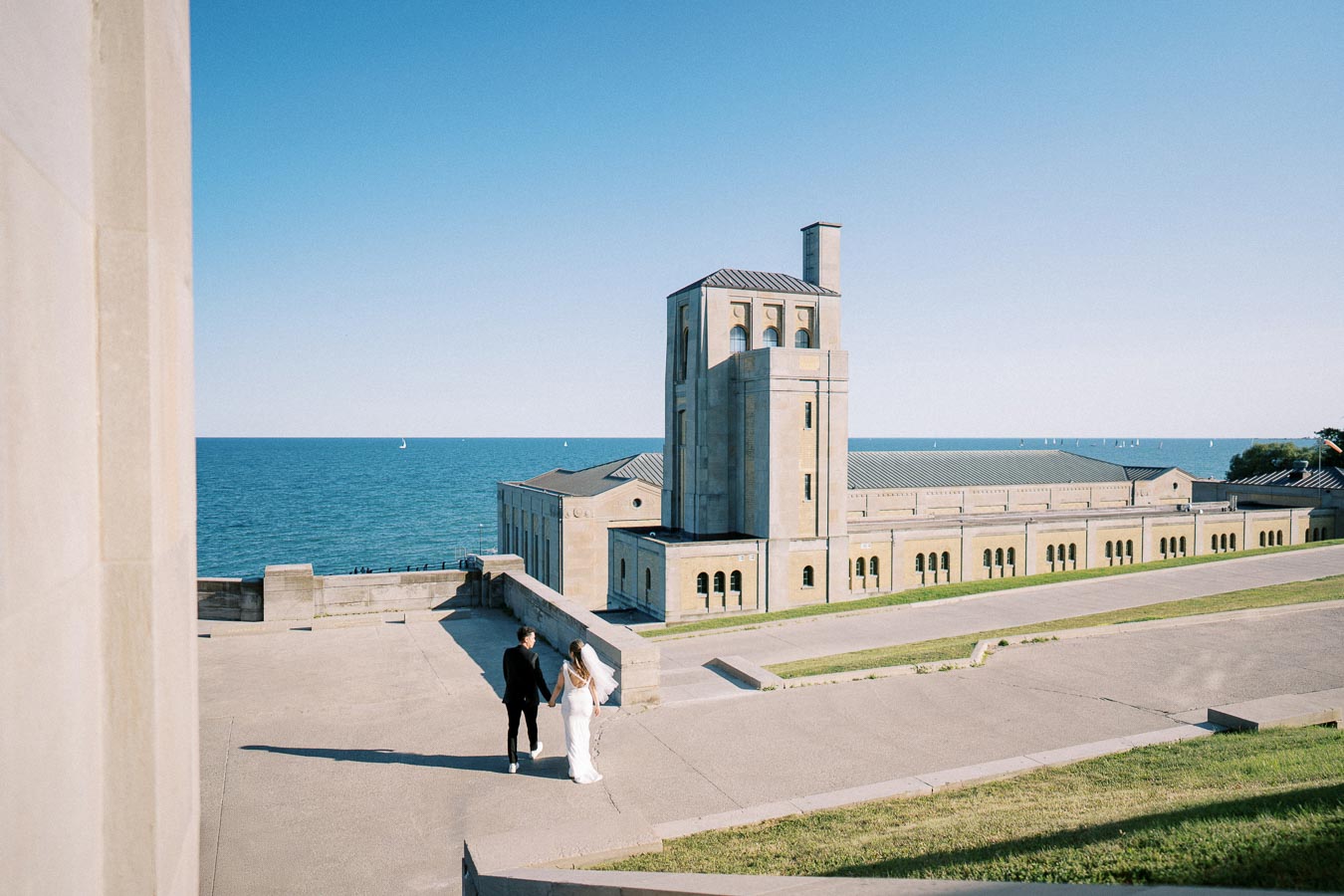 A couple walks hand in hand near a historic stone building by the sea on a sunny day, with a clear blue sky and distant sailboats in the background.
