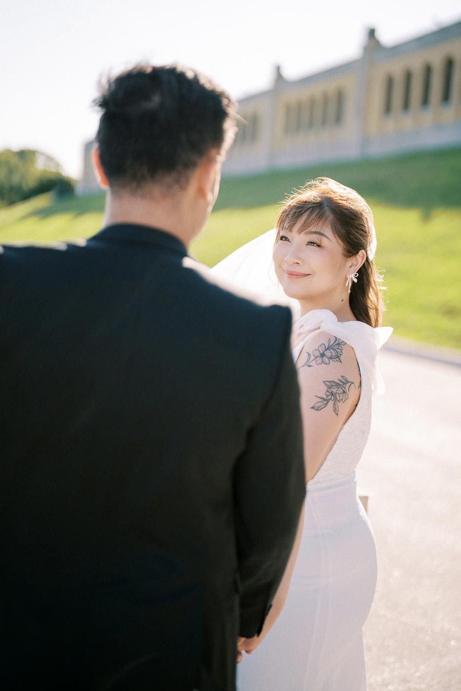 A bride in a white dress with floral tattoos on her shoulder smiles at a groom in a black suit, standing outdoors on a sunny day with a green landscape and building in the background.