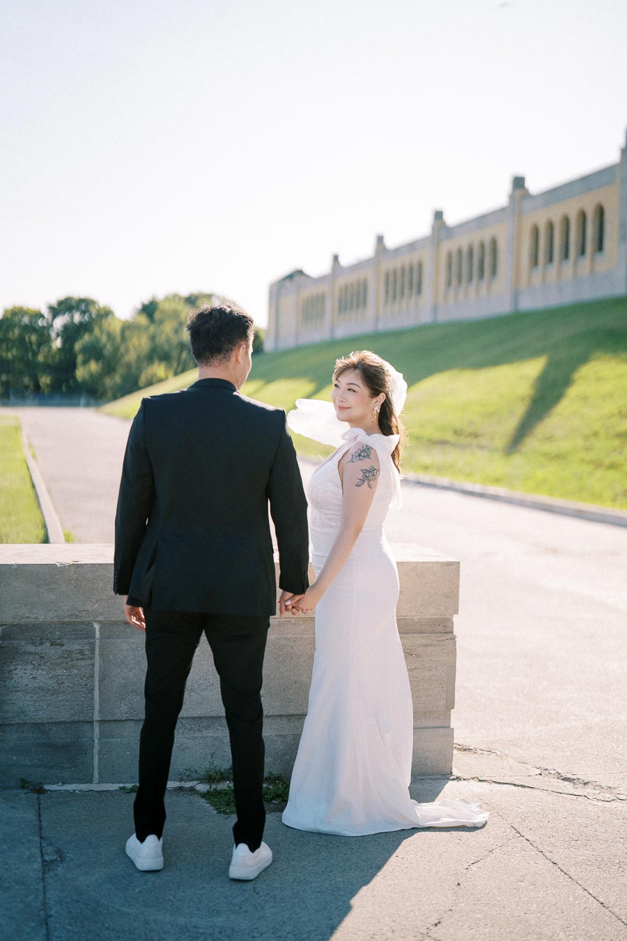 Bride and groom holding hands outdoors, with elegant architecture and greenery in the background on a sunny day.