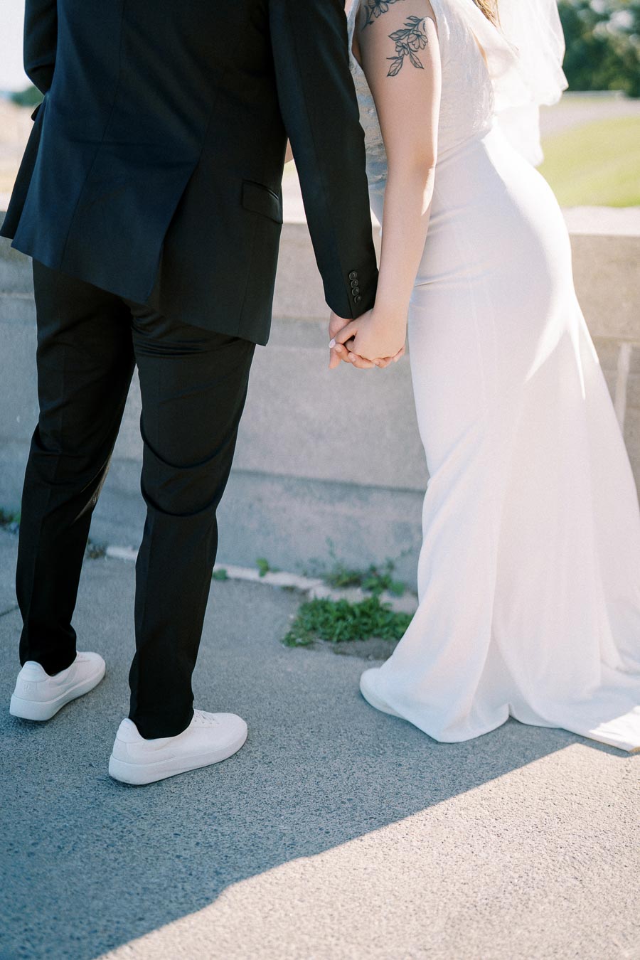 A couple holding hands in wedding attire, the bride wearing a white gown and the groom in a black suit with matching sneakers, standing outdoors.