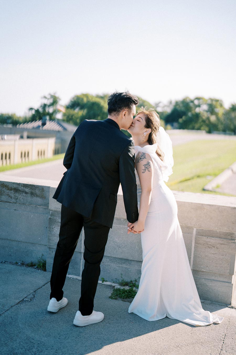 A bride and groom share a romantic kiss on their wedding day, standing by a stone wall in a picturesque outdoor setting. The bride is wearing a white gown with a train, while the groom is dressed in a black suit and white sneakers. They hold hands, symbolizing love and commitment.