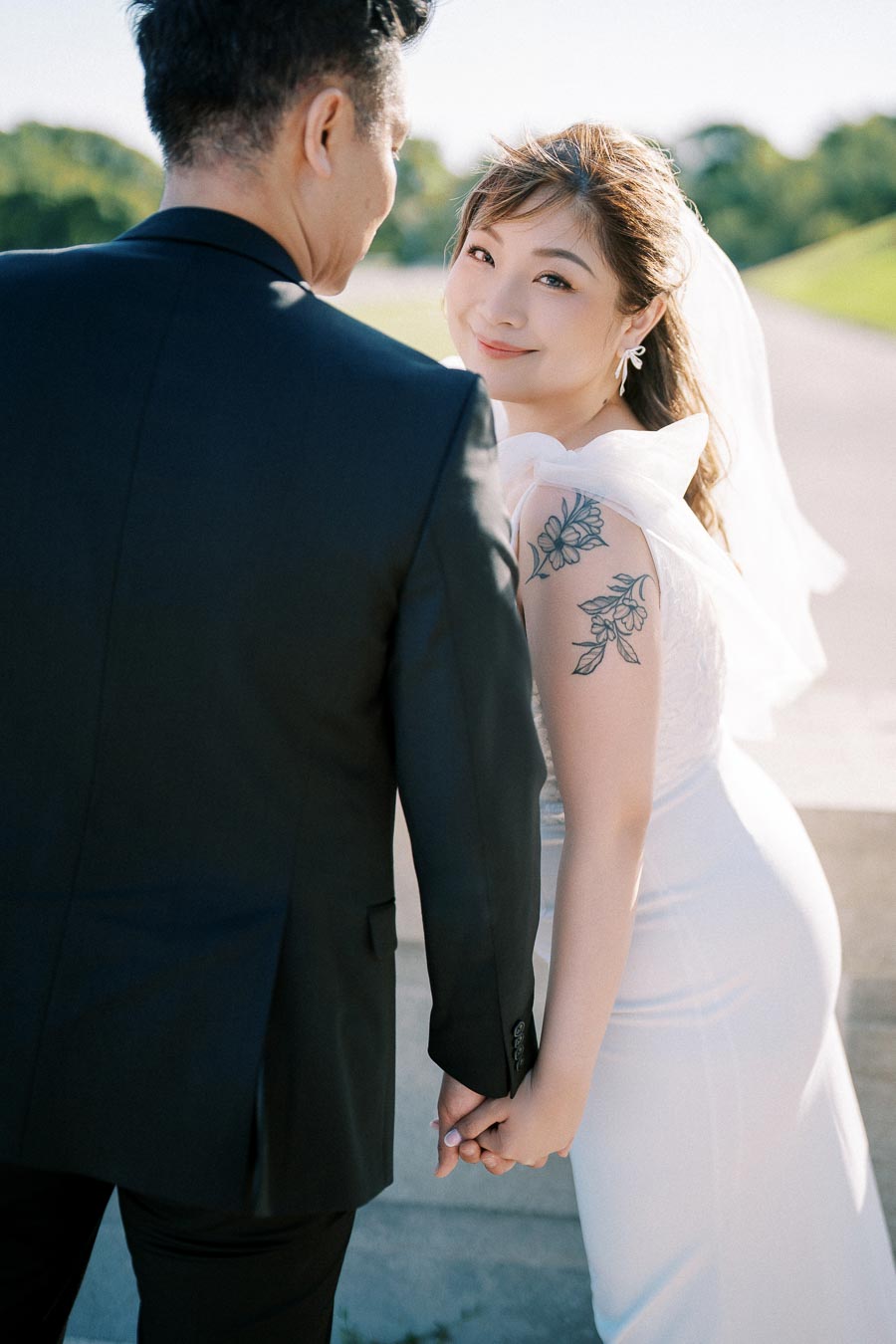 Bride and groom holding hands during an outdoor wedding, with the bride smiling over her shoulder and sunlight illuminating her floral tattoo and white dress.