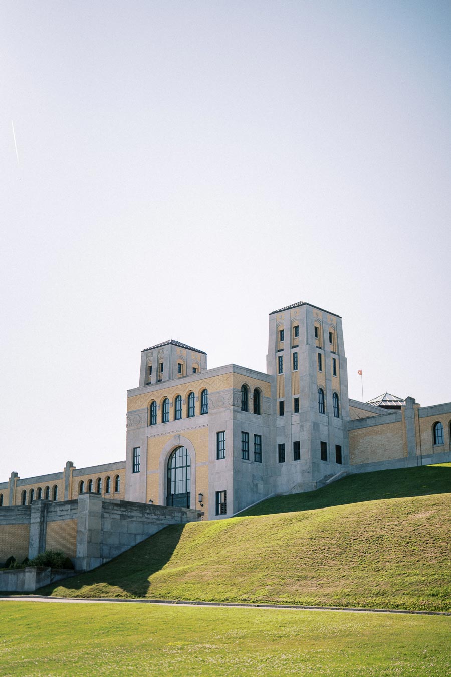 Historic building with towers and large arched windows set on a grassy hill under a clear blue sky.