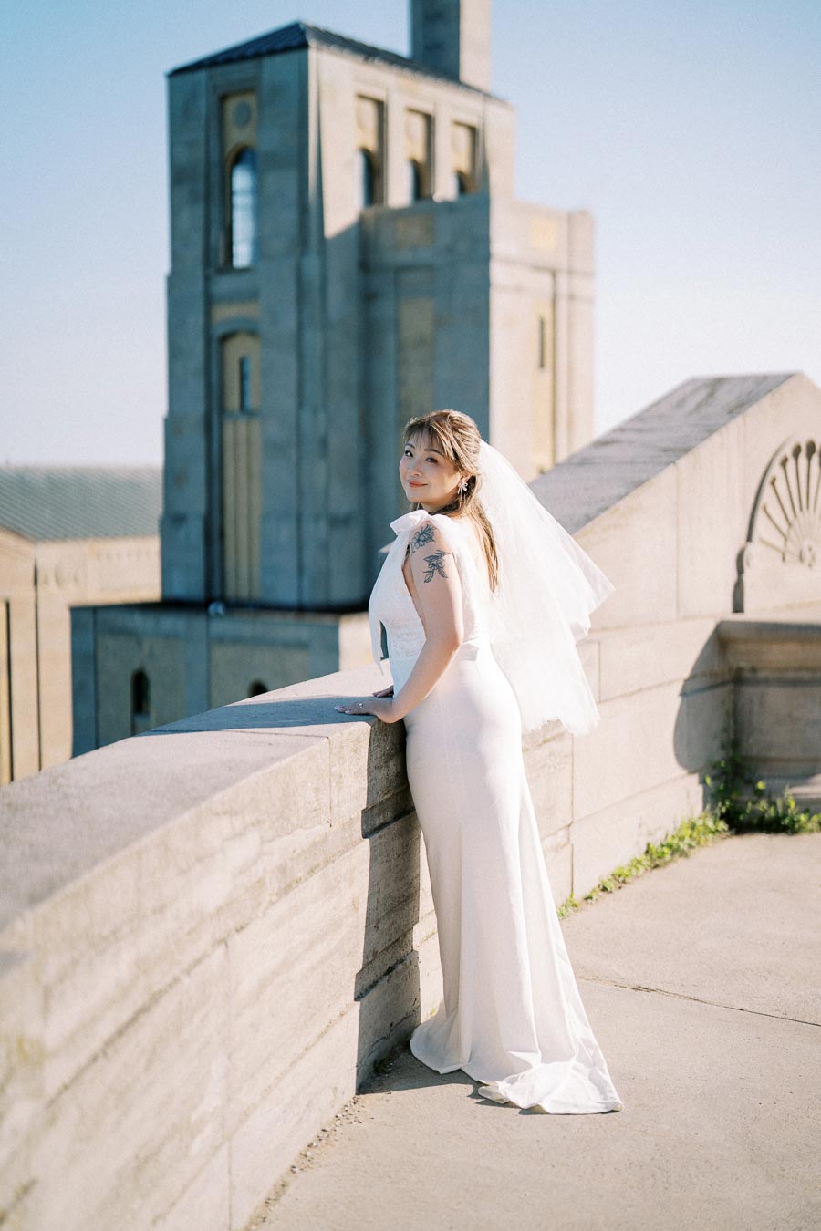 Elegant bride in a white wedding dress and veil posing on a sunlit stone terrace with historic architecture background.