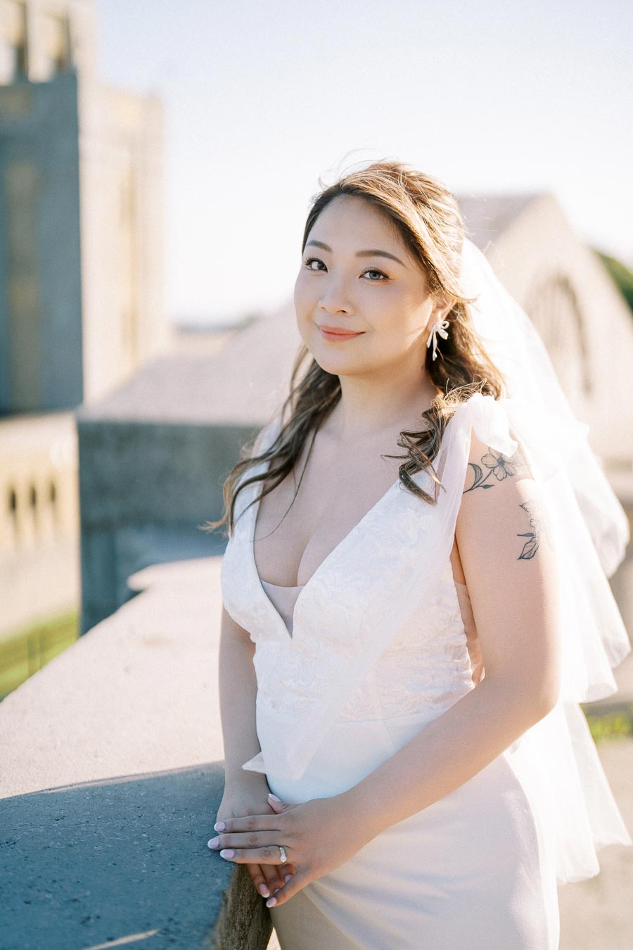 A smiling bride in an elegant white wedding dress and veil, standing on a sunlit balcony, with a historic building in the background.