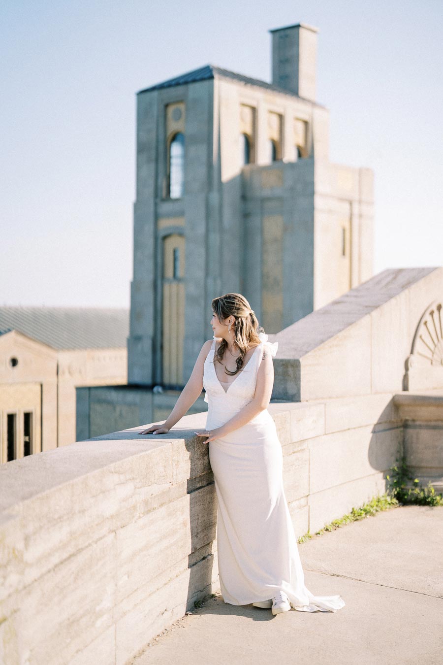 Bride in elegant white gown standing on a historic stone terrace, admiring the architectural beauty of a vintage building under a clear blue sky.