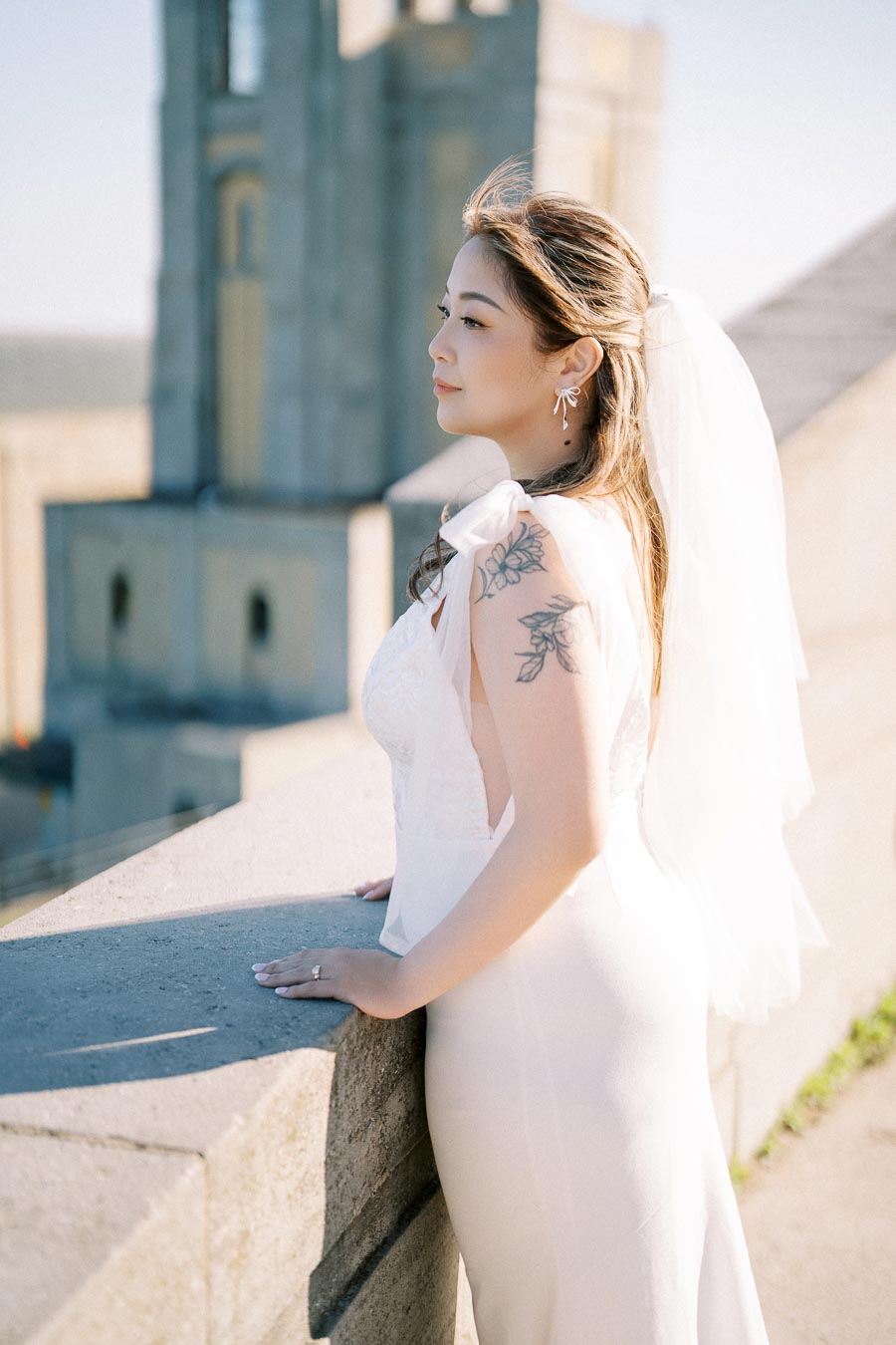 A bride in a white wedding dress with a floral tattoo on her arm stands on a stone terrace, gazing thoughtfully into the distance, with a blurred historic building in the background.