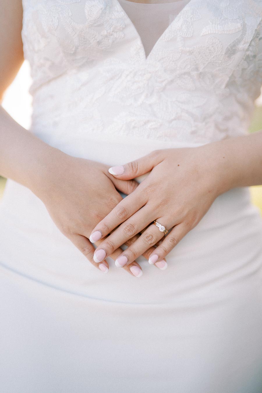 Close-up of a bride's hands gracefully crossed over a white wedding dress, showcasing a diamond engagement ring and manicured nails with a lace bodice detail.