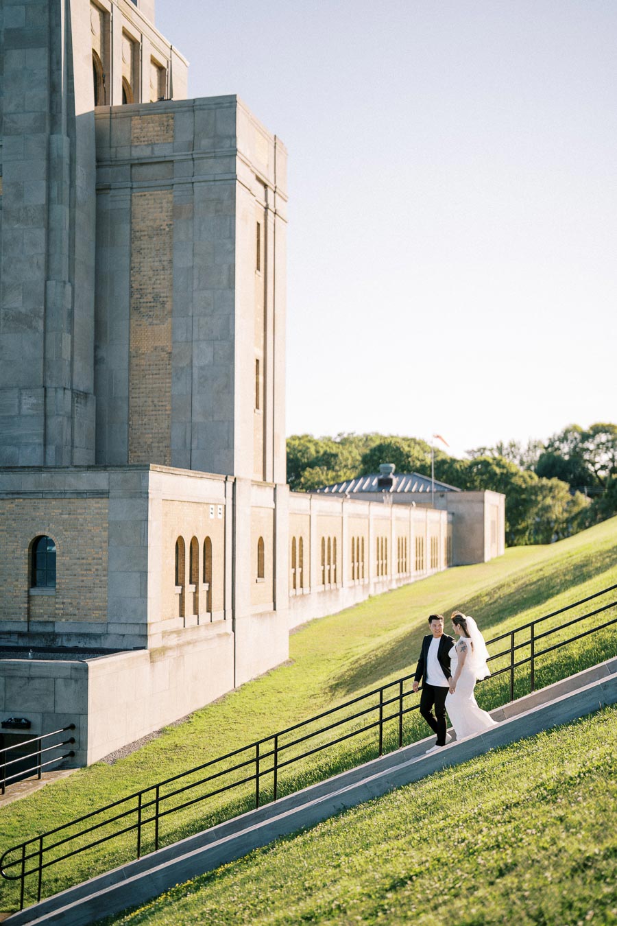Wedding couple walking down a grassy hill beside a historic building, bathed in natural sunlight.