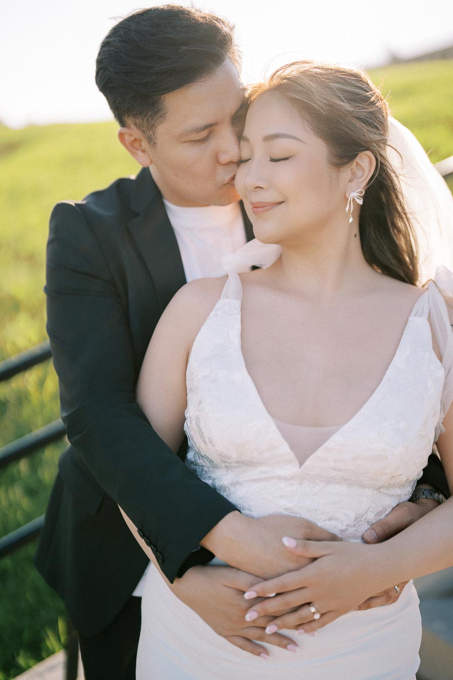 Bride and groom embracing outdoors with a serene, sunny backdrop, highlighting their smiling faces and wedding attire