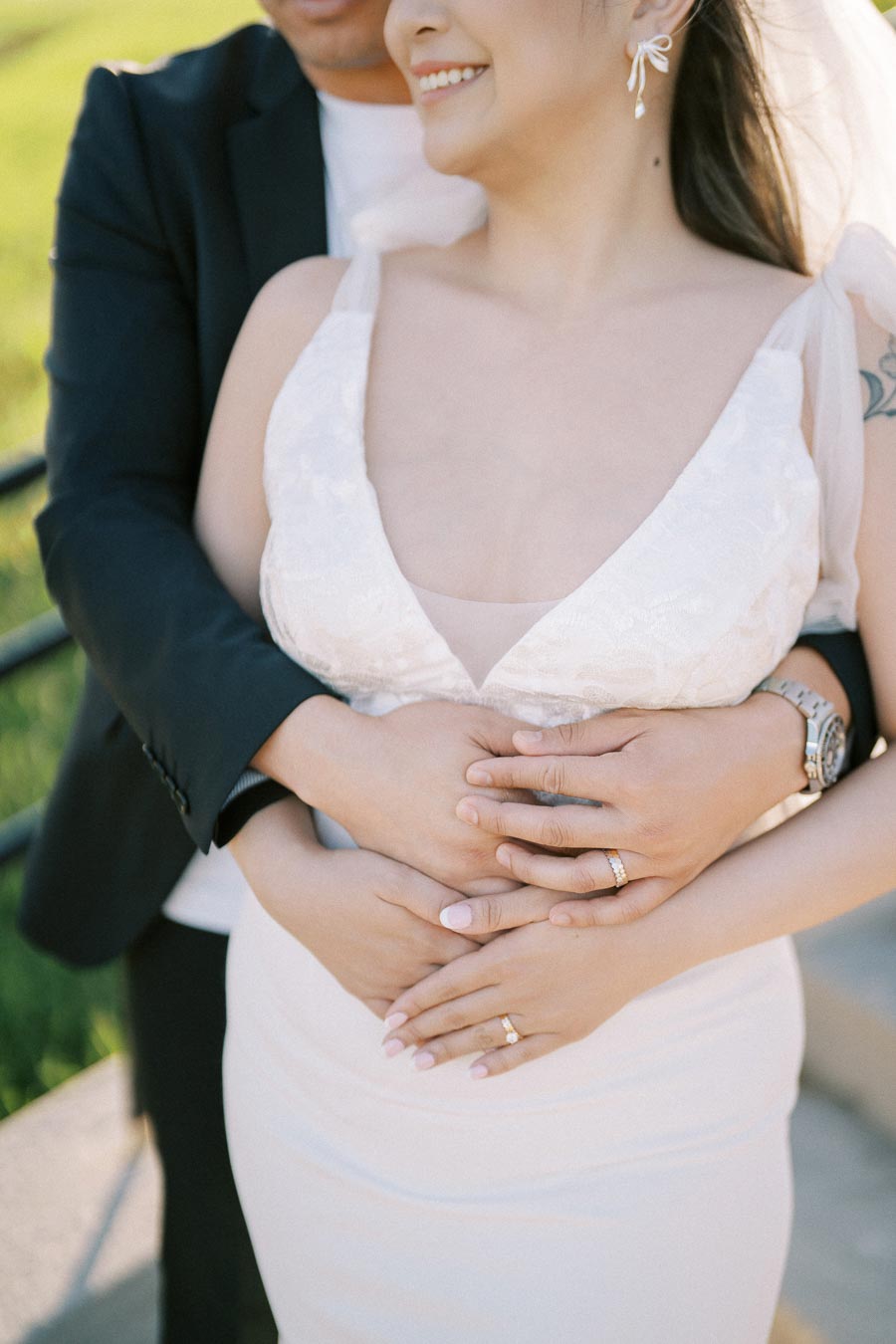 Close-up of a couple embracing, highlighting wedding rings on their hands, set against a sunny outdoor backdrop.