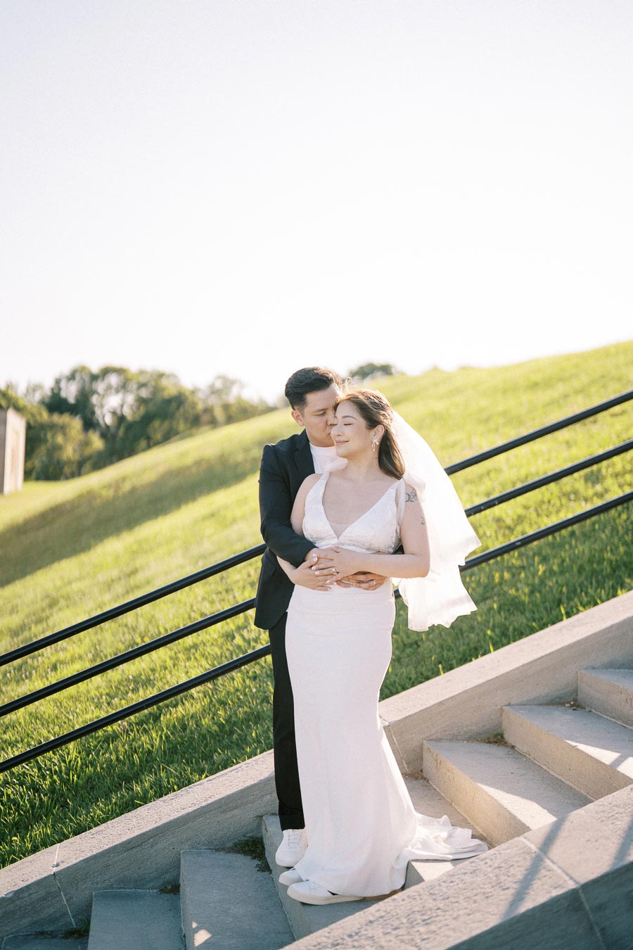 A bride and groom embrace on a sunlit outdoor staircase, with lush green grass and a clear blue sky in the background, capturing a romantic wedding moment.