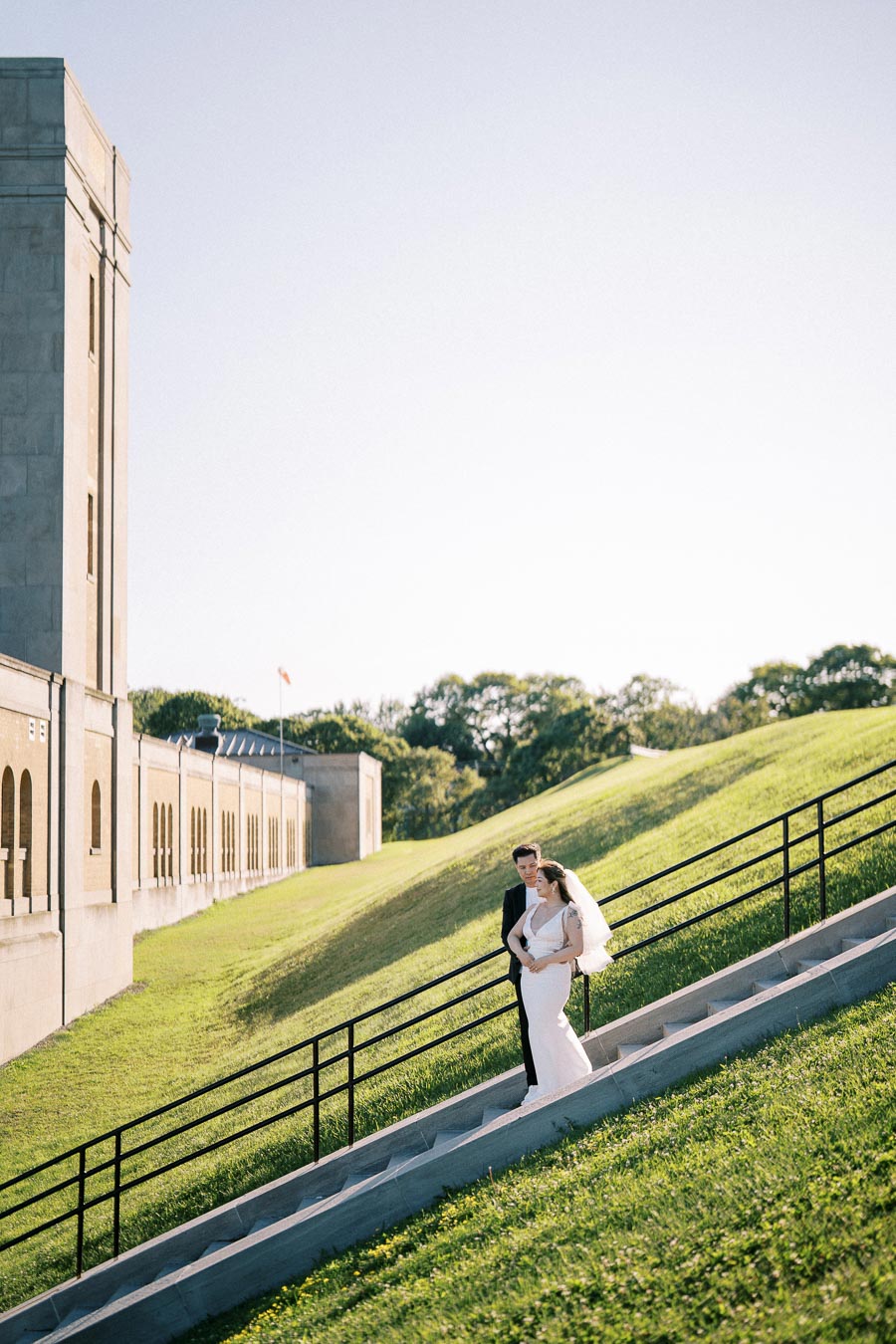 Bride and groom walking down stairs in elegant wedding attire beside a historic stone building on a sunny day.