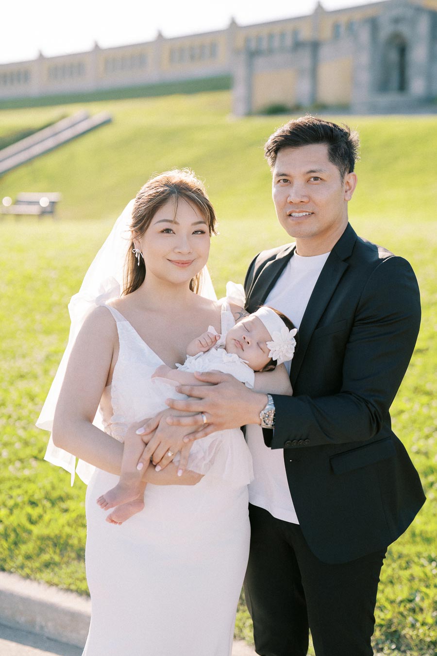 A happy family posing outdoors on a sunny day, featuring a mother in a white dress holding a baby, and a father in a black jacket.