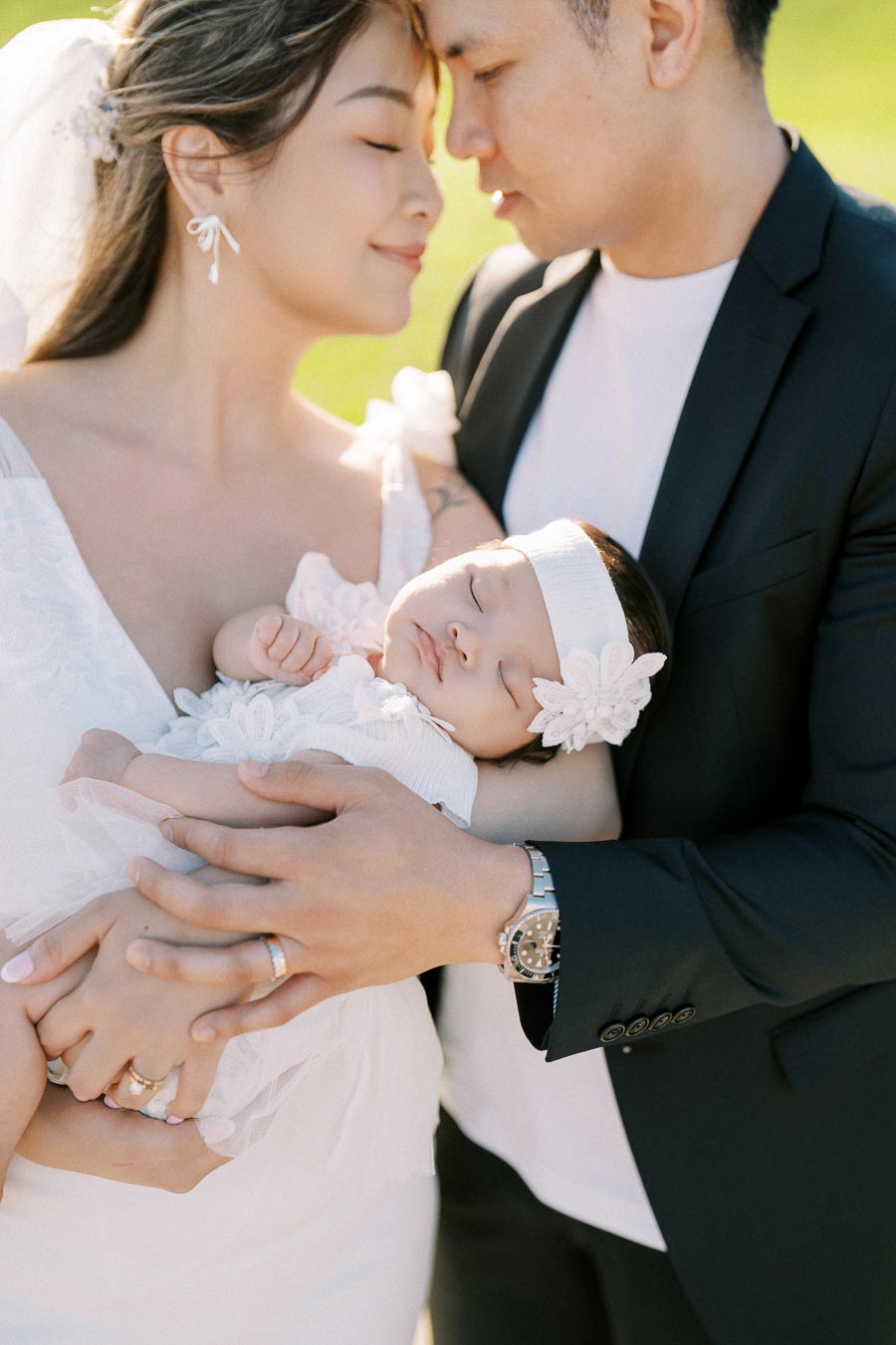 A happy couple lovingly embraces their sleeping baby, all dressed in white, standing together in a serene outdoor setting.