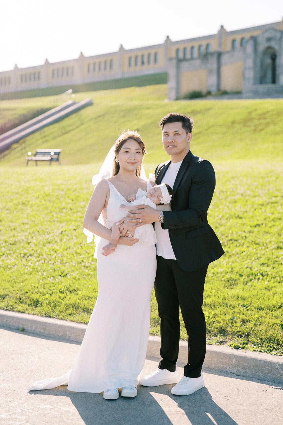 A couple in wedding attire with a baby, standing on a sunny day in front of a grassy hill and large building.