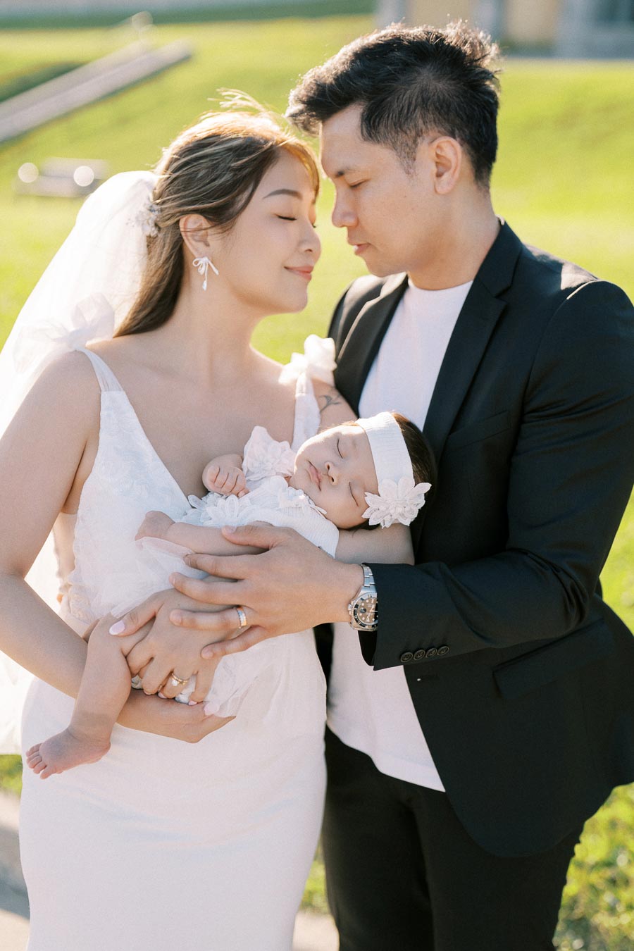 A loving couple holding their sleeping baby in an outdoor setting, with the mother in a white dress and veil, and the father in a black suit, showcasing family bonding and serene moments.