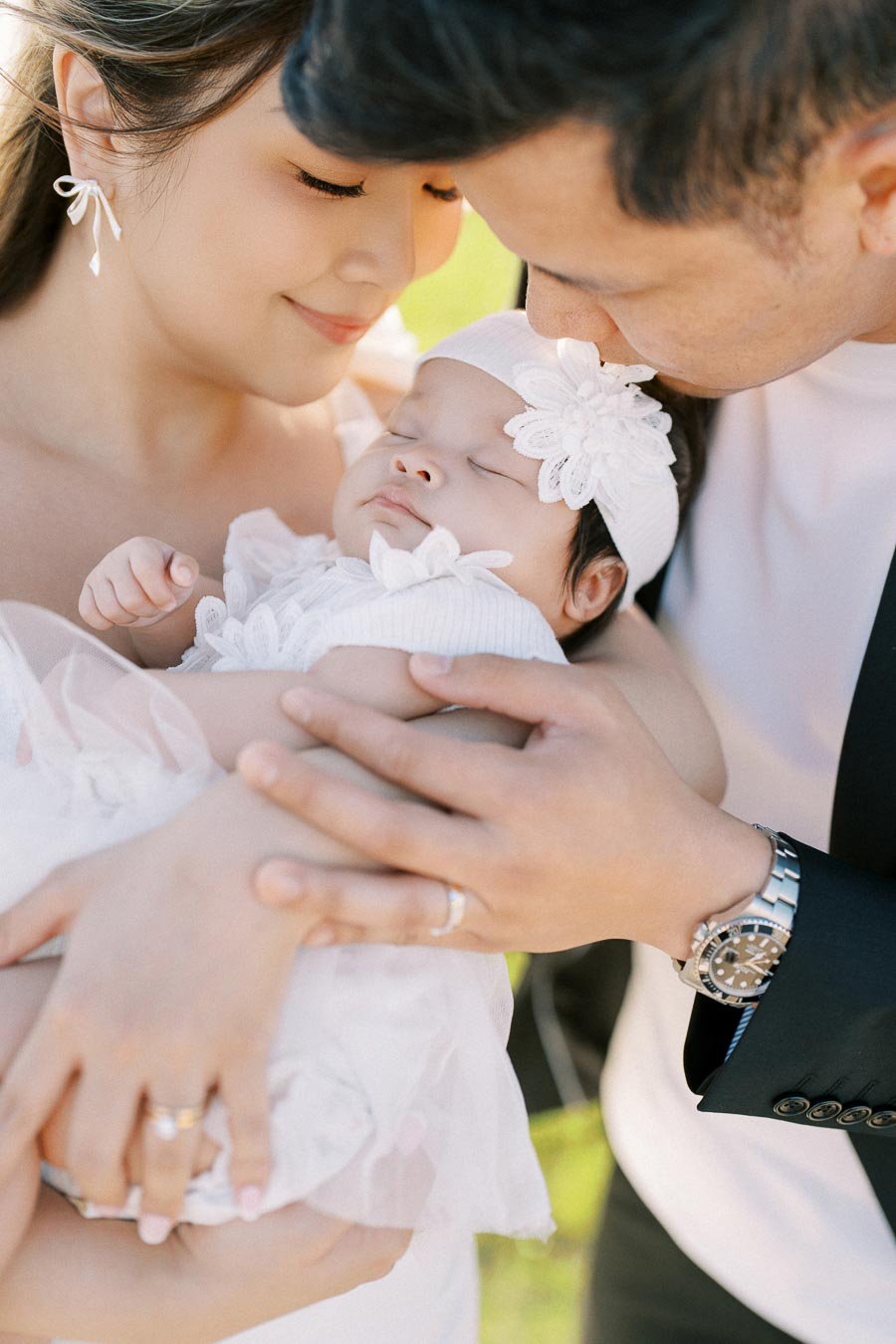 Happy family bonding with parents lovingly holding their sleeping baby dressed in white, featuring delicate floral headband, in a serene outdoor setting.