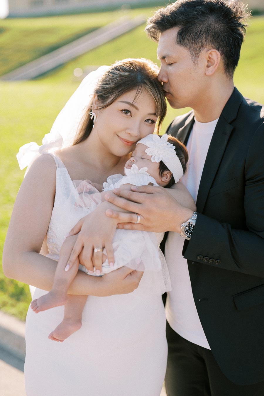 A loving couple dressed elegantly, with the man gently kissing the woman's forehead as she holds a sleeping baby adorned with a flower headband, all standing in a sunlit outdoor setting.