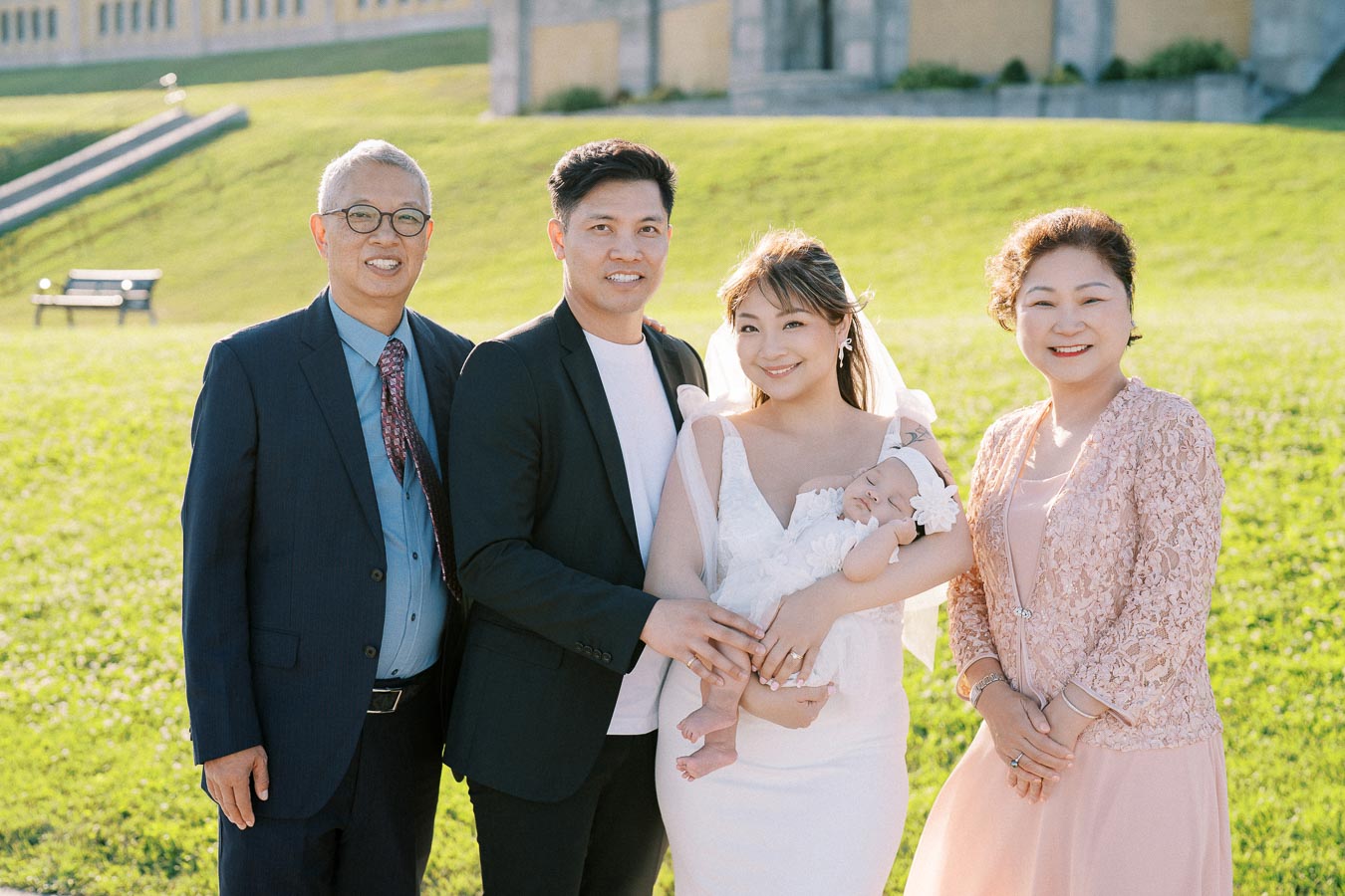 A happy family posing outdoors on a sunny day, featuring a couple holding a baby and standing alongside two smiling elders, all dressed in formal attire.