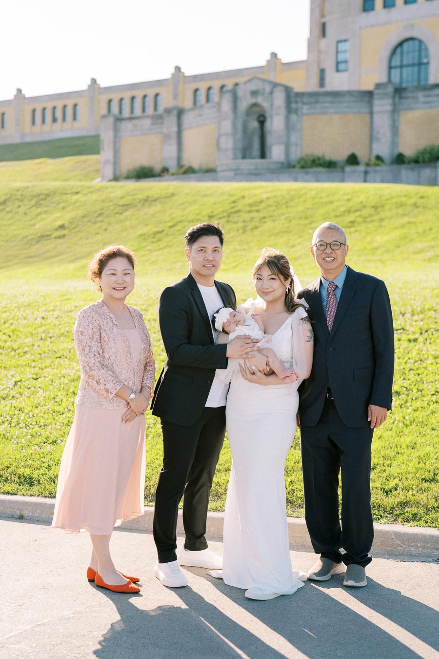 A family standing together on a sunny day, with adults and a baby dressed in elegant clothes, posing in front of a large historic building on a grassy hill.
