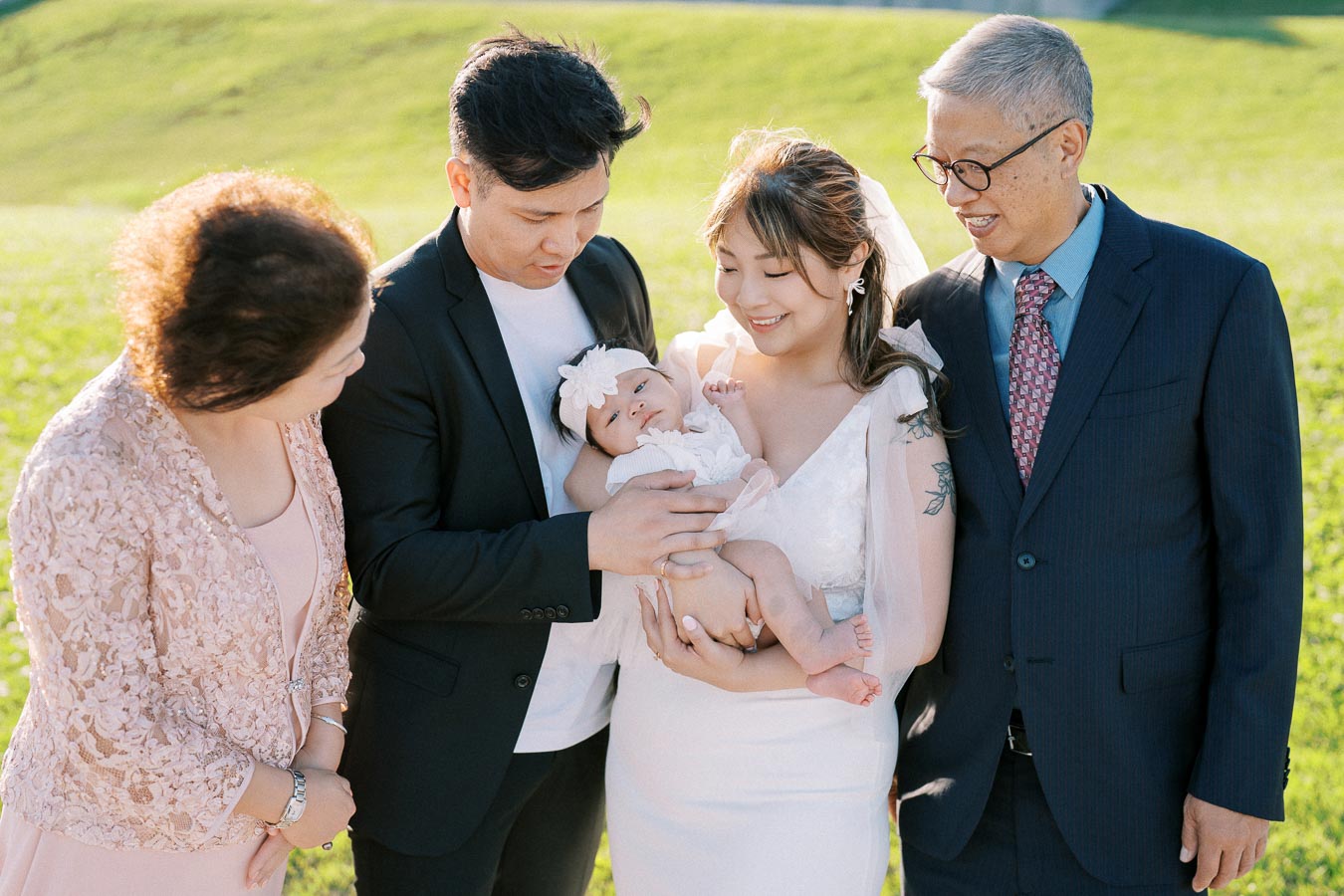 A family gathered outdoors, smiling and admiring a baby being held by its mother. The group stands on a lush green field under bright sunlight.