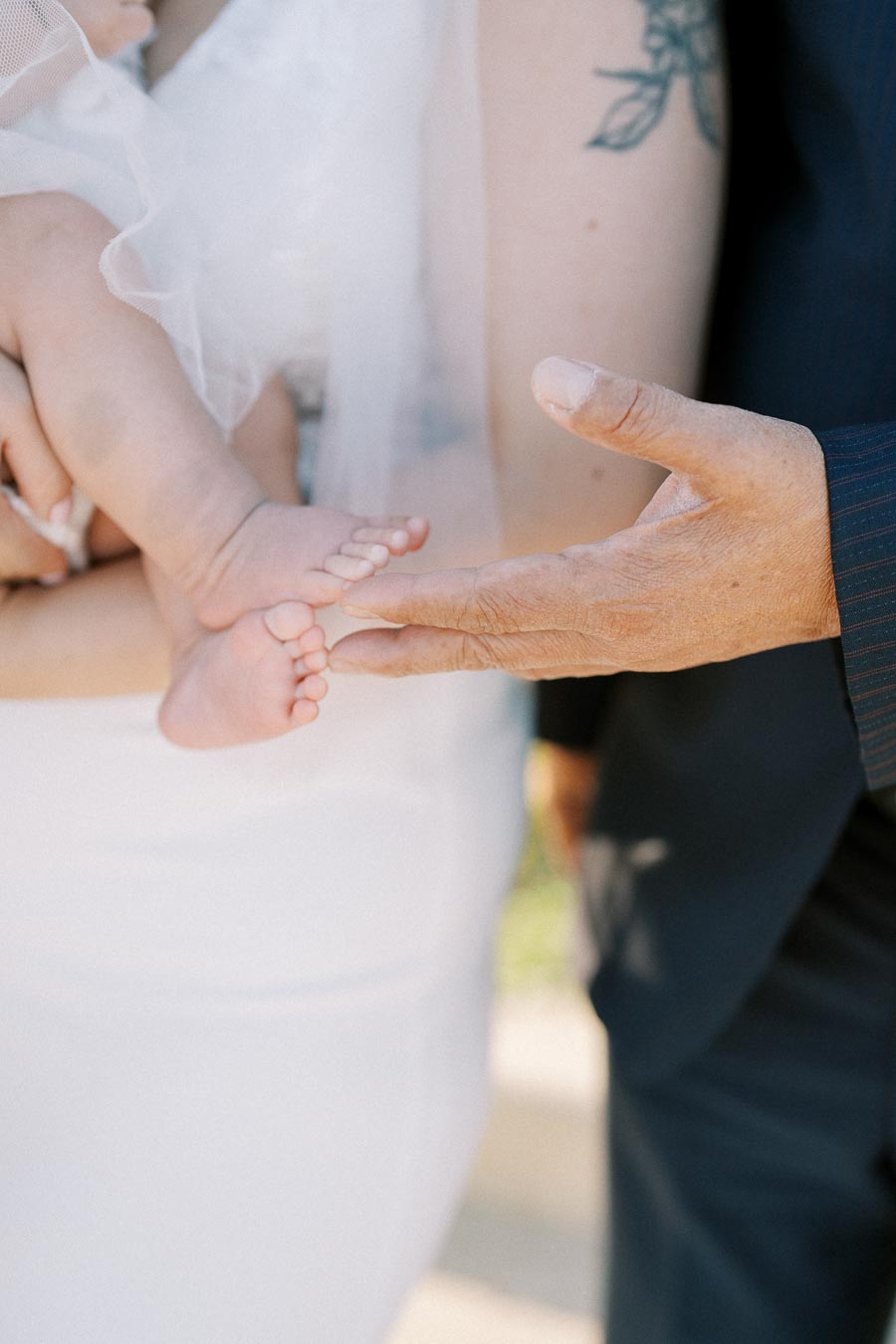 Close-up of an adult gently holding the small feet of a baby, showcasing tenderness and connection, with a wedding dress and tattoo visible in the background.