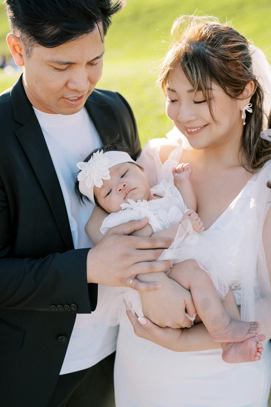 A loving family moment captured outdoors, with a couple holding their baby dressed in white, surrounded by greenery and smiling warmly.