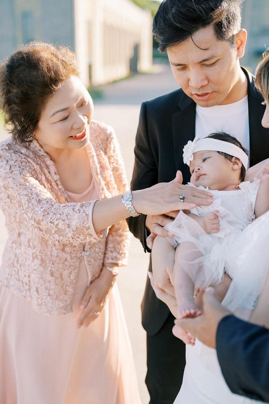 A joyful family moment featuring a stylishly dressed woman in a pink lace outfit smiling warmly at a baby girl in a delicate white dress and headband, held by a man in a black suit.