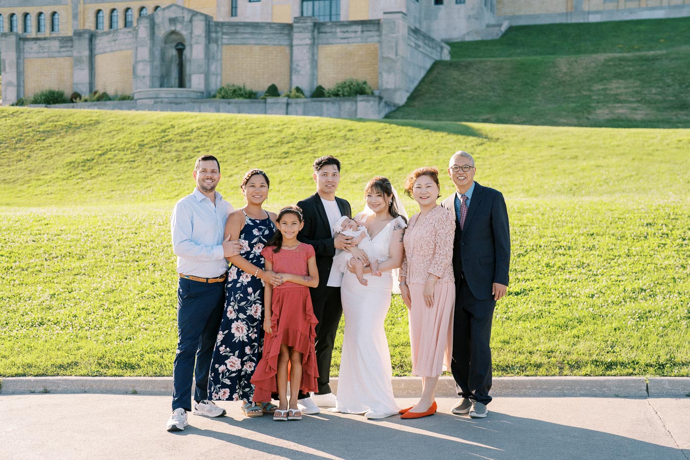 A family group photo standing outside in a sunny setting with a historic yellow brick building in the background. The group includes adults, a child, and a baby, all smiling and dressed in semi-formal attire.