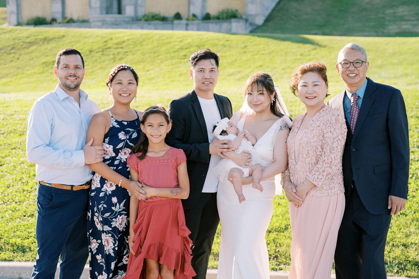 A happy, multi-generational family gathers for a group photo outdoors on a sunny day, with members dressed in formal and semi-formal attire, including a bride holding a baby, surrounded by lush green grass.