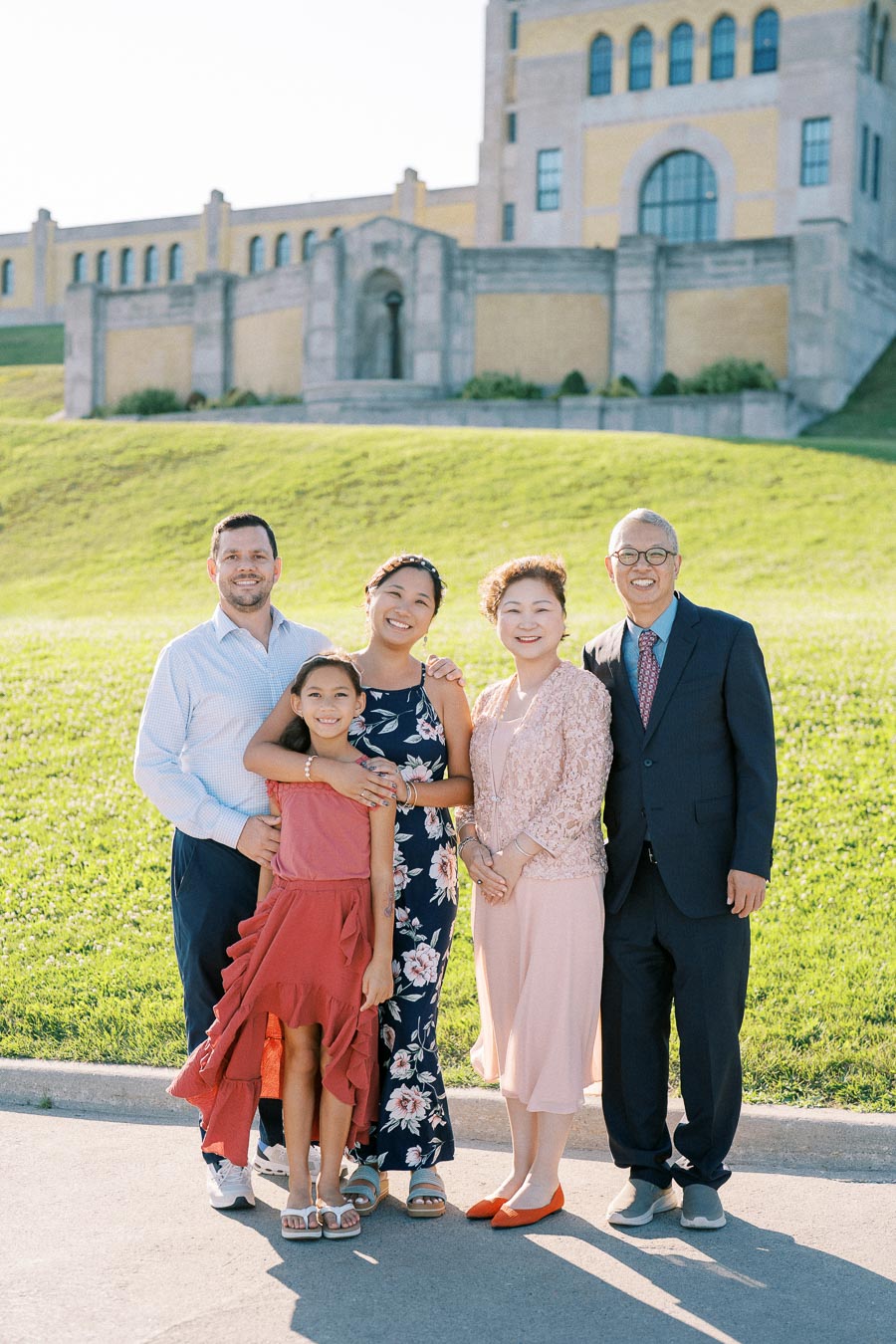 A diverse family standing outside on a sunny day, in front of a historic building with green lawns, all smiling and dressed in colorful, formal attire.