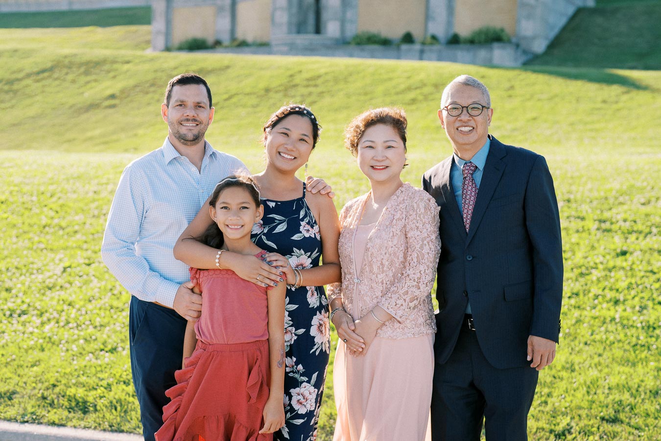 A cheerful multigenerational family standing together on a sunny day with a green lawn background, displaying diverse attire and smiles.
