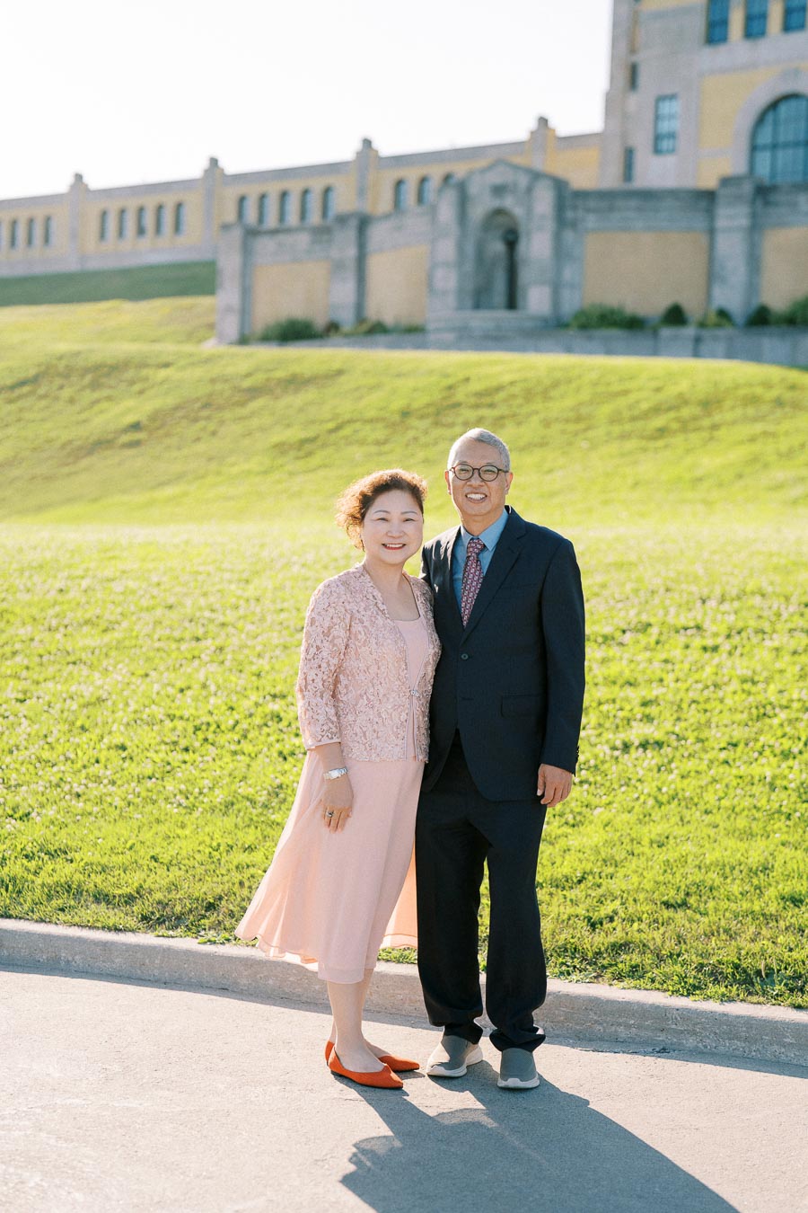 Smiling couple standing together outdoors in front of a historic building, wearing elegant attire on a sunny day, with a grassy hill in the background.